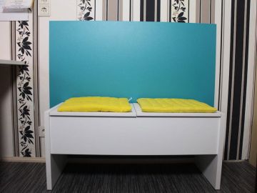 Weiße Sitzbank mit zwei gelben Sitzkissen vor einem türkisfarbenen Wandschutz; im Hintergrund Tapete mit schwarzen und beigefarbenen Streifen und Blumenmustern.