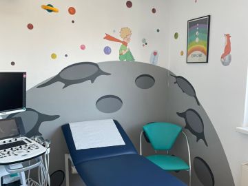 Kinderfreundlich gestalteter Untersuchungsraum mit Ultraschallgerät, Liege und türkisfarbenm Stuhl; an der Wand ein großes Wandbild aus Wandpaneelen mit Motiven aus „Der kleine Prinz“, inklusive Planet, Prinz, Fuchs und farbiger Planeten.