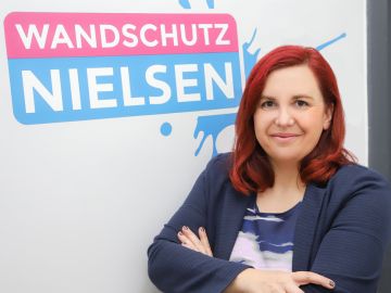 Geschäftsführerin Nancy Nielsen steht mit verschränkten Armen vor einer Wand mit dem Firmenlogo „Wandschutz Nielsen“.