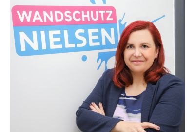 Geschäftsführerin Nancy Nielsen steht mit verschränkten Armen vor einer Wand mit dem Firmenlogo „Wandschutz Nielsen“.
