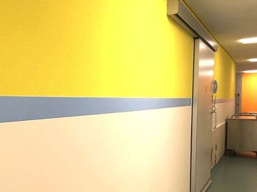 Krankenhausflur mit gelber Wandfarbe, abgetrennt durch einen grauen Streifen aus einem Wandpaneel
