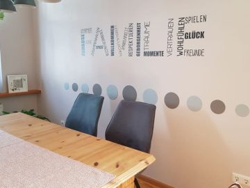 Essbereich mit hellem Holztisch, zwei grauen Polsterstühlen und einer Wandgestaltung mit bunten Punkten und Schriftzügen wie „Zuhause“, „Vertrauen“, „Glück“ und „Wohlfühlen“.