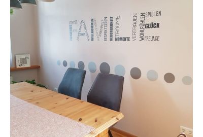 Essbereich mit hellem Holztisch, zwei grauen Polsterstühlen und einer Wandgestaltung mit bunten Punkten und Schriftzügen wie „Zuhause“, „Vertrauen“, „Glück“ und „Wohlfühlen“.