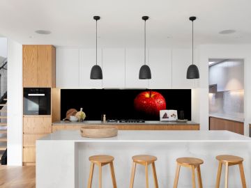 Moderne, offene Küche mit weißer Kochinsel, Holz- und Weißfronten, vier Holzbarhockern und schwarzen Hängelampen; auffällig ist die schwarze Küchenrückwand mit einem großformatigen Fotodruck eines roten Apfels mit Wassertropfen.