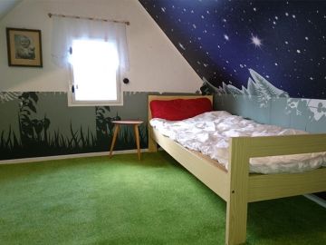 Gemütliches Kinderzimmer mit Einzelbett, grünem Teppich und fantasievoller Wandgestaltung: eine Wand zeigt eine Wiese in Silhouettenoptik, die Dachschräge ein Sternenhimmel-Motiv.
