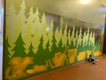 Wandgestaltung in einem Kindergartenflur mit stilisierten grünen Tannenbäumen und gelben Tierillustrationen am unteren Rand, darunter Mäuse und weitere Waldtiere.
