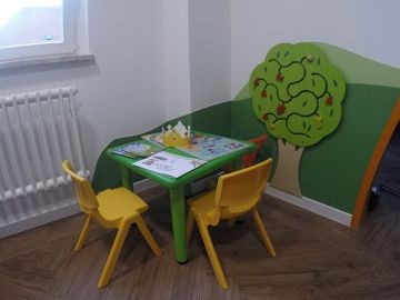 Bunte Kinderspielecke mit grünem Tisch, zwei gelben Stühlen und einer Wanddekoration in Form eines grünen Baums mit beweglichen Elementen. Auf dem Tisch liegen Ausmalbilder, eine Papierkrone und Kinderhefte.