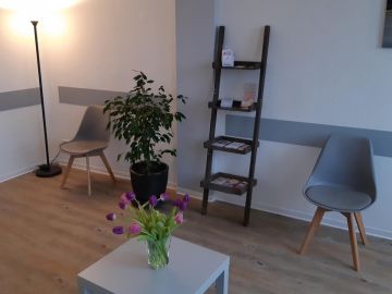 Modern eingerichtetes Wartezimmer mit grauen Stühlen, einer schwarzen Stehlampe, einem kleinen Regal in Leiterform mit Infomaterial. Weiße Wände mit grauen Stuhllehnenschutz und gleichzeitig Dekoleiste.