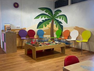 Freundlich gestaltete Kinderspielecke einer Arztpraxis mit bunten Stühlen an der Wand, großem Spieltisch mit Bausteinen und Wandpaneelen in Form einer Palme und Wüstenlandschaft.