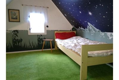Gemütliches Kinderzimmer mit Einzelbett, grünem Teppich und fantasievoller Wandgestaltung: eine Wand zeigt eine Wiese in Silhouettenoptik, die Dachschräge ein Sternenhimmel-Motiv.
