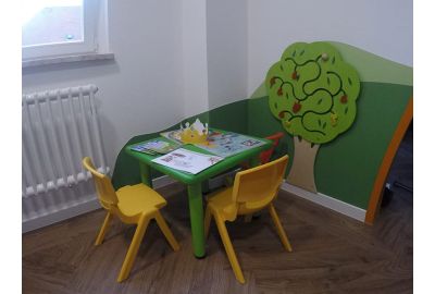 Bunte Kinderspielecke mit grünem Tisch, zwei gelben Stühlen und einer Wanddekoration in Form eines grünen Baums mit beweglichen Elementen. Auf dem Tisch liegen Ausmalbilder, eine Papierkrone und Kinderhefte.