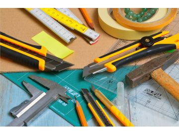 Verschiedene Bastel- und Handwerksmaterialien auf einem Arbeitstisch, darunter Cutter-Messer, Hammer, Maßband, Zirkel, Schneidematten, Lineale, Klebeband, Bleistifte und ein Messschieber.